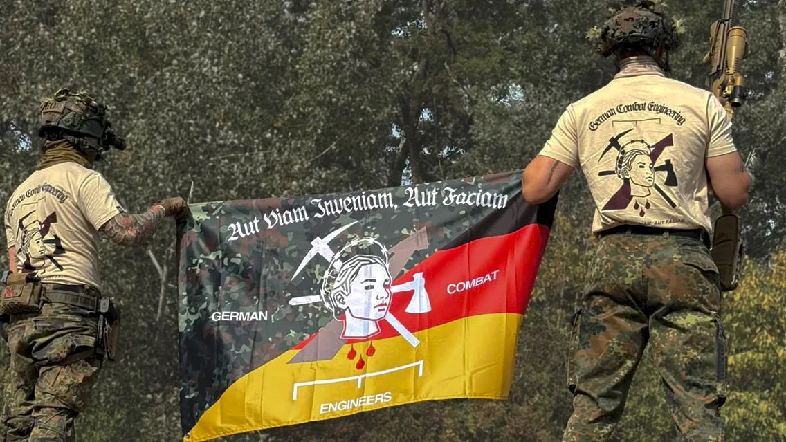 Zwei Männer in Flecktarn und Gefechtshelm halten von hinten gesehen die Pionier Flagge, beide tragen das Pionier Shirt