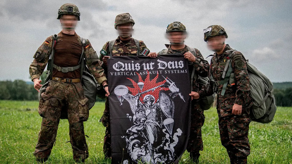 Vier Fallschirmjäger mit gepackten Sprungsäcken halten auf einer Wiese die Quis-ut-Deus-Flagge mit dem Erzengel Michael