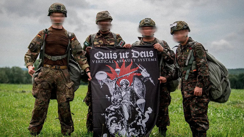 Vier Fallschirmjäger mit gepackten Sprungsäcken halten auf einer Wiese die Quis-ut-Deus-Flagge mit dem Erzengel Michael