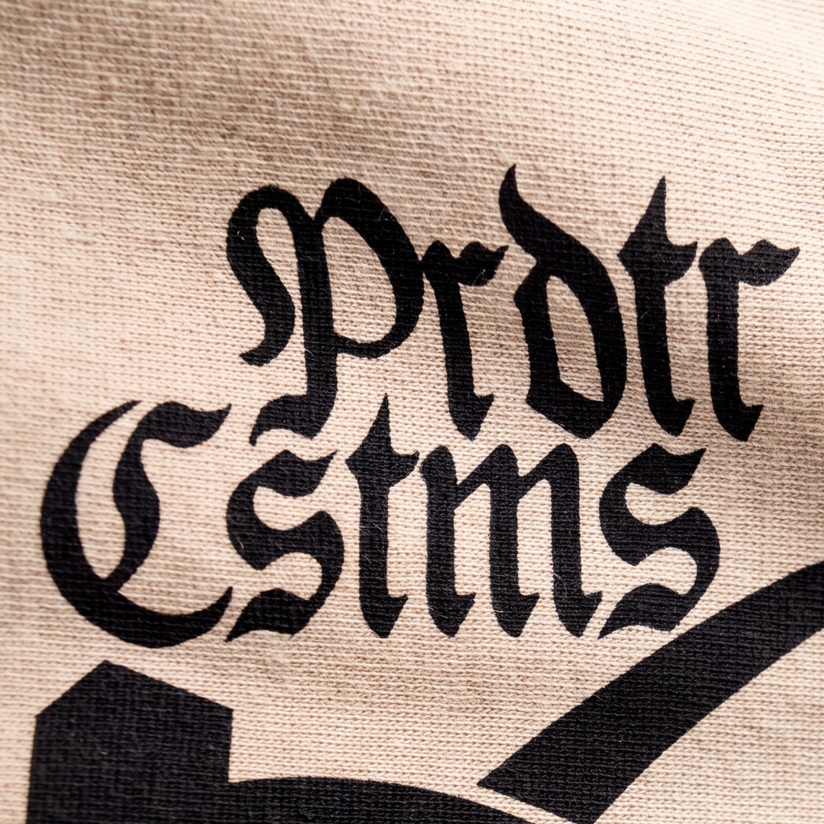 Detailaufnahme des Brustdrucks vom Pionier Shirt: der Schriftzug Prdtr Cstms in gebrochener Schrift, schwarz auf sandfarbenem Baumwolljersey, darunter die Oberkante des gedruckten Zeichens. Die Maschen des Stoffs sind durch die Druckfarbe sichtbar.